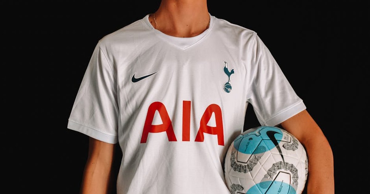 Adult soccer player in Tottenham Hotspur jersey holding a ball against a dark background.