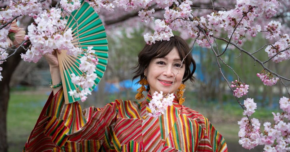 Woman in vibrant kimono holding a fan amidst cherry blossoms in spring.