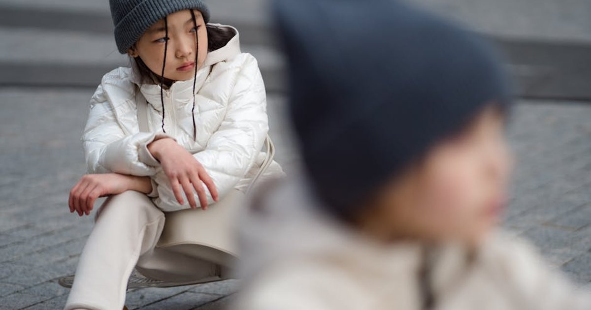 A young girl wearing a bonnet and white jacket sits on a skateboard on concrete pavement.