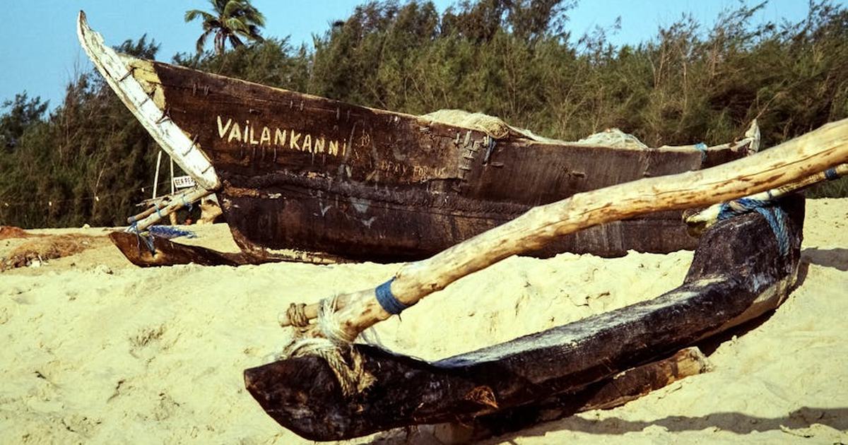 A wooden fishing boat named 'Vailankanni' rests on a sandy beach in India, surrounded by lush trees.
