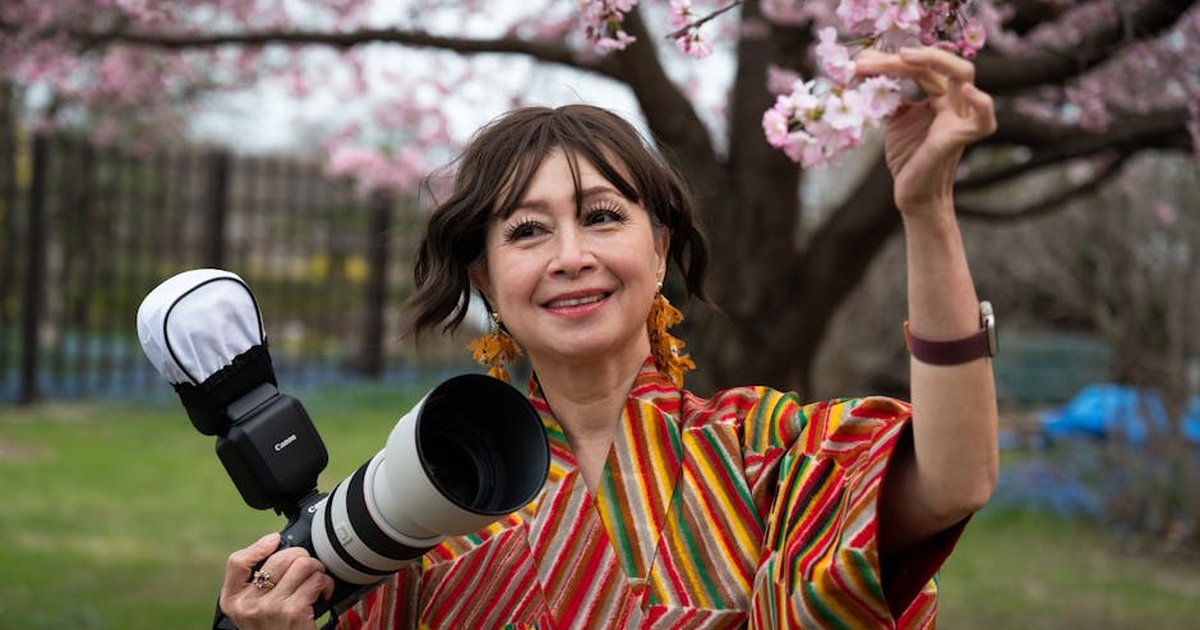 Smiling woman in a colorful kimono holds a camera under cherry blossoms, capturing spring beauty.
