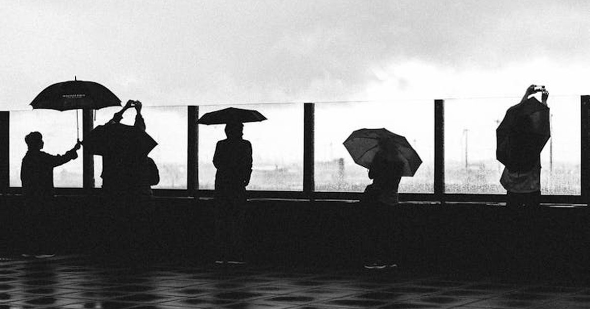 Black and white image of people holding umbrellas on a rainy day, capturing silhouettes against a moody sky.