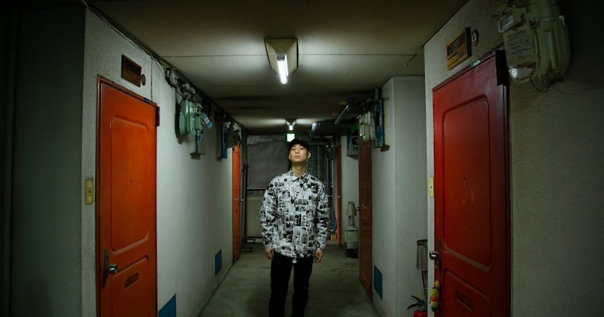 Full body of Asian male wearing stylish clothes standing in narrow corridor with doors in apartment house