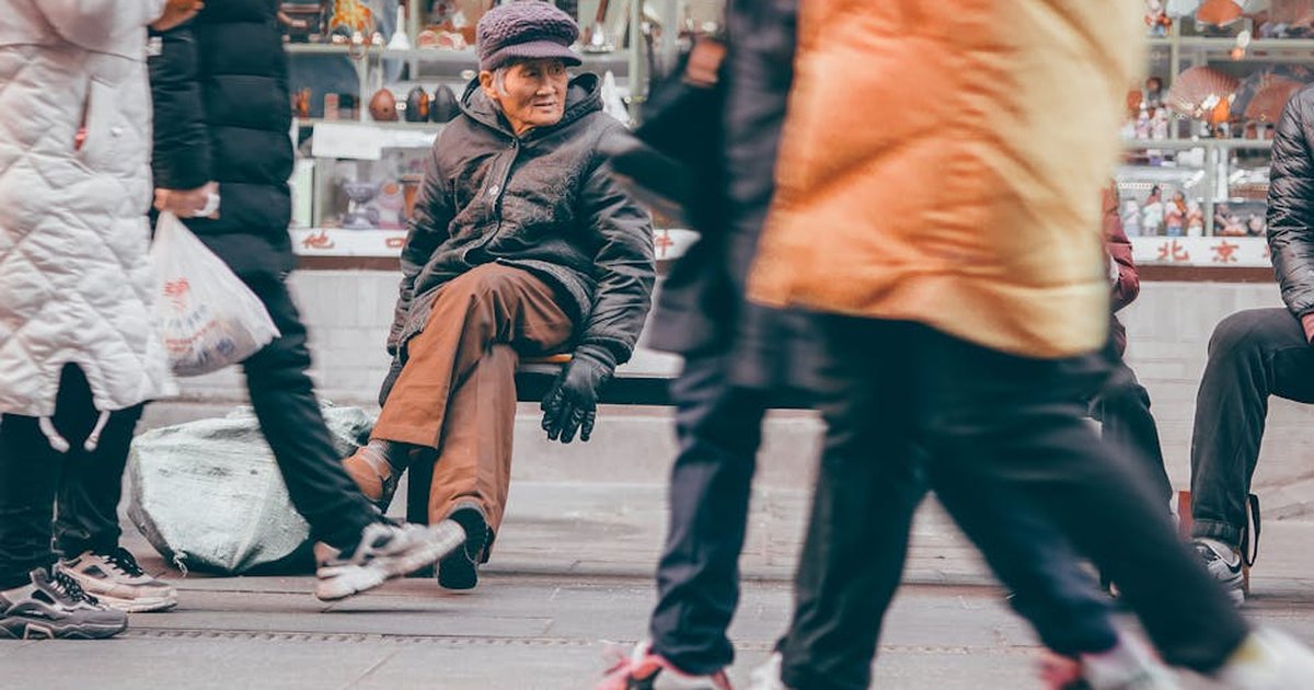 A candid street scene in Beijing featuring an elderly man sitting on a bench amid bustling pedestrians.