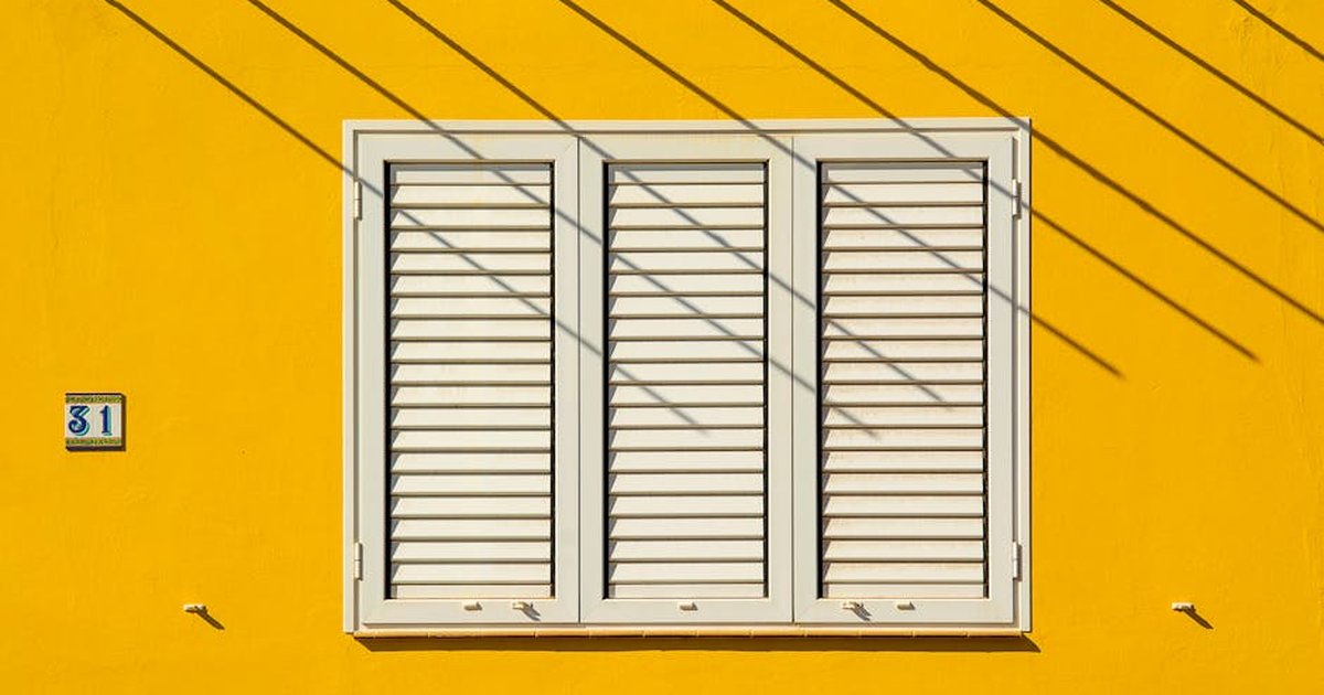 White shuttered window on a vibrant yellow wall with geometric shadows. House number 31 visible.