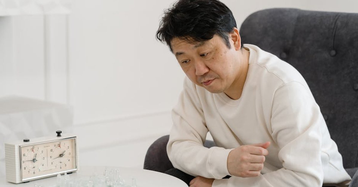 Asian man contemplating a chess game indoors, wearing a white sweater, with an analog clock nearby.