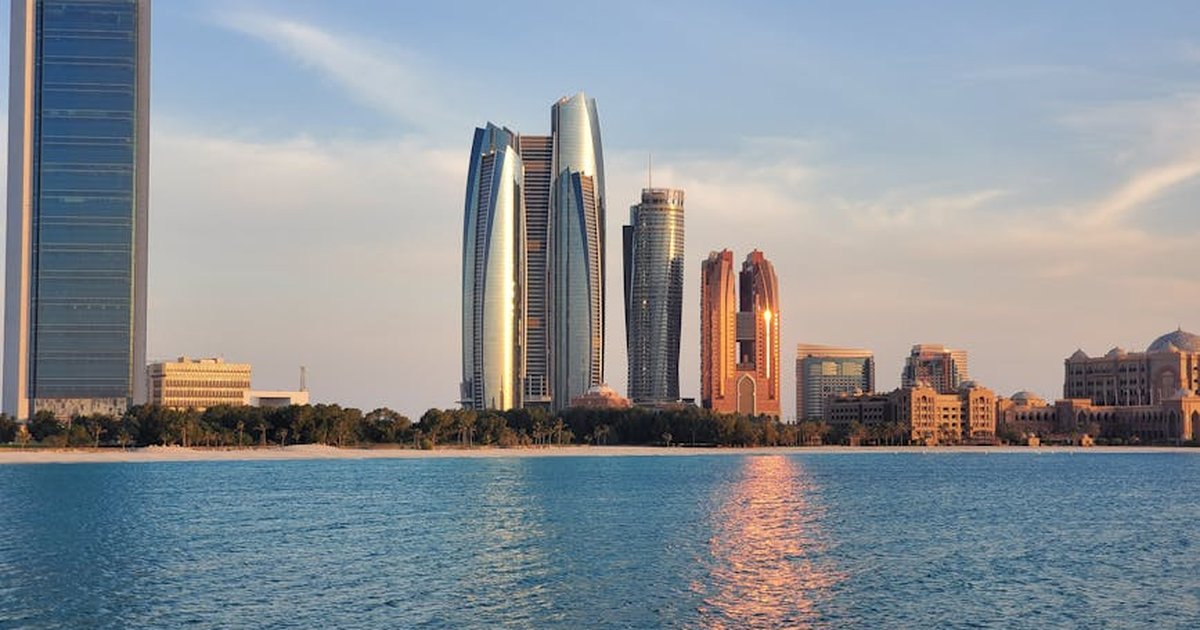 Capture of Abu Dhabi skyline with modern skyscrapers reflecting on calm sea at sunset.