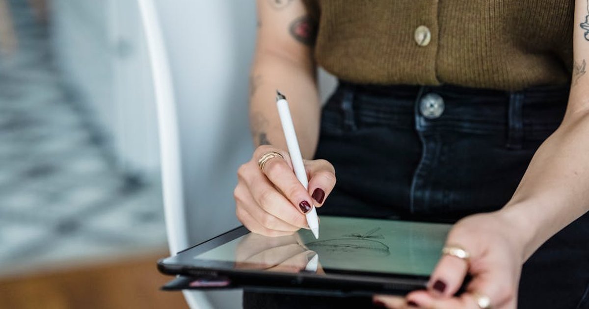 Crop anonymous lady in stylish clothes drawing on graphic tablet with stylus pen while sitting in chair in bright apartment
