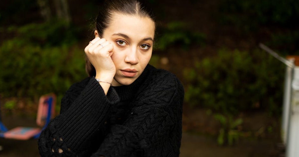 Young woman in a black sweater with hand on cheek, outdoor setting, soft lighting.
