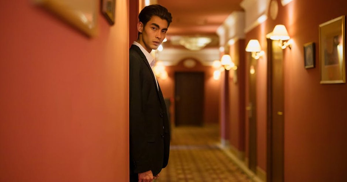 A fashionable man in a sleek black suit poses confidently in an elegant hotel hallway.