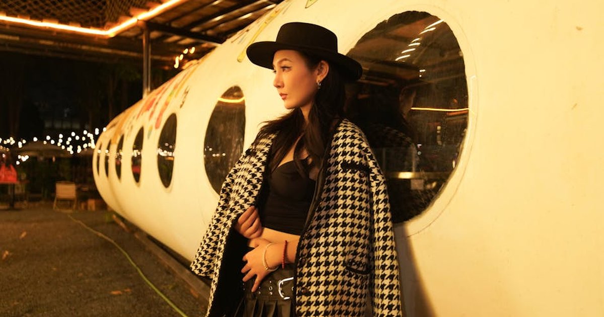 A fashionable woman in a houndstooth coat leans against a tube-shaped structure, illuminated by night lights.