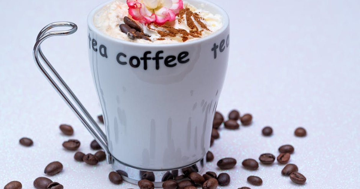 A stylish coffee mug with whipped cream, cinnamon, a pink flower, and scattered coffee beans on a white background.