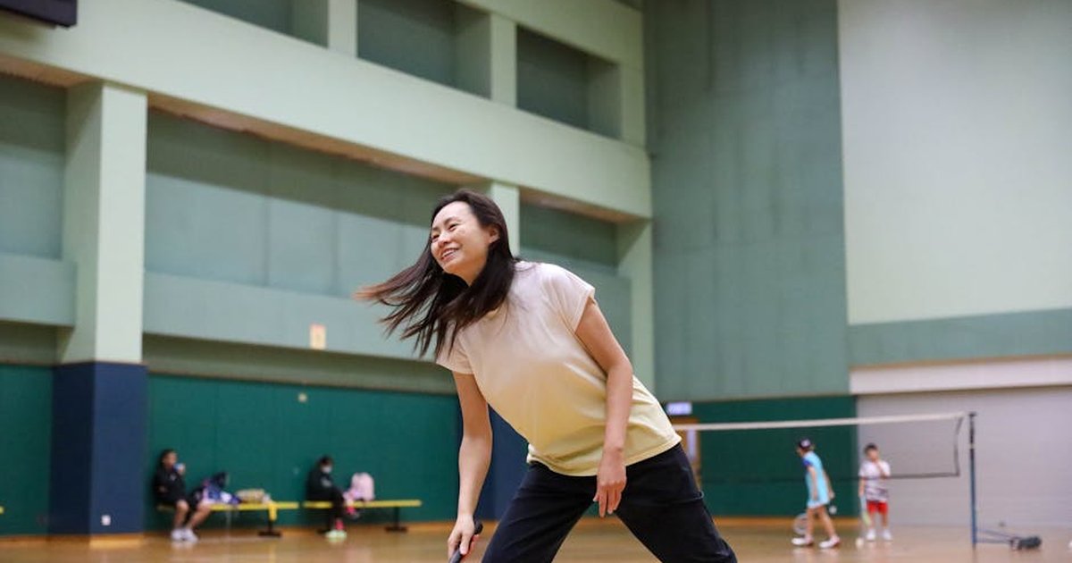 Smiling Asian woman playing badminton indoors in a sports hall, wearing casual sportswear.