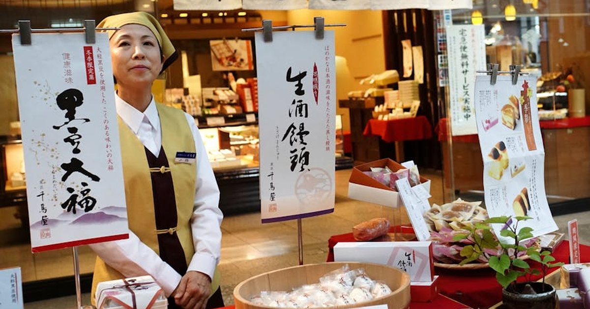 Japanese woman presenting traditional sweets at a storefront with signage. Authentic cultural setting.