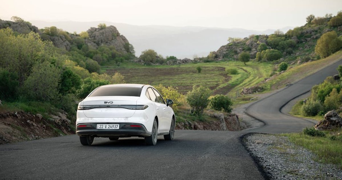 A white car drives on a winding road through scenic hills in Shaqlawa, Iraq during daylight.
