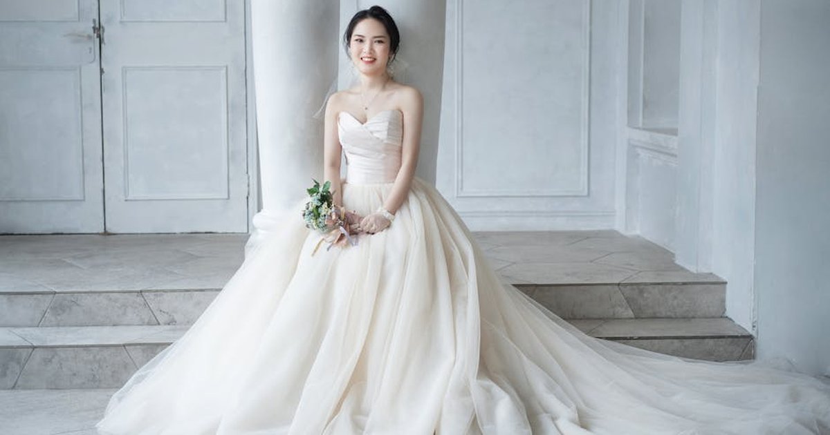 Beautiful bride seated indoors in a flowing wedding gown holding a small bouquet.