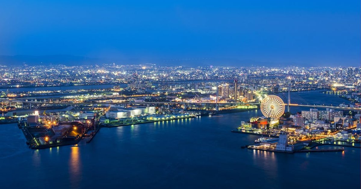 Aerial view of a vibrant city skyline by night featuring a glowing Ferris wheel and illuminated waterfront.