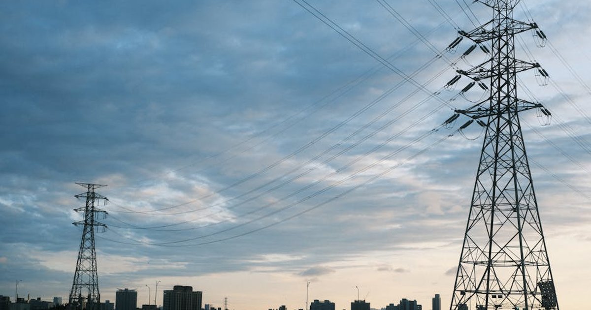 A city skyline at dusk with transmission towers and power lines silhouetted against a cloudy sky.