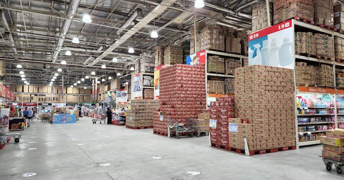 A large wholesale warehouse interior filled with stacked merchandise and shoppers browsing the aisles.