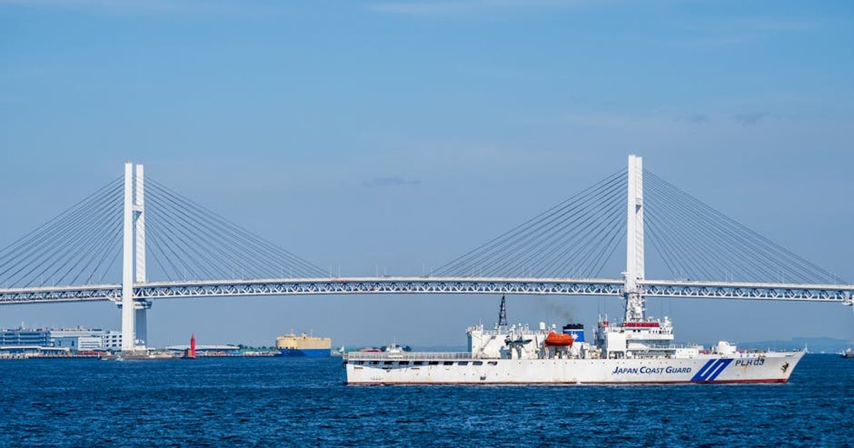 Japan Coast Guard vessel sailing in front of the iconic Yokohama Bay Bridge under a clear blue sky.