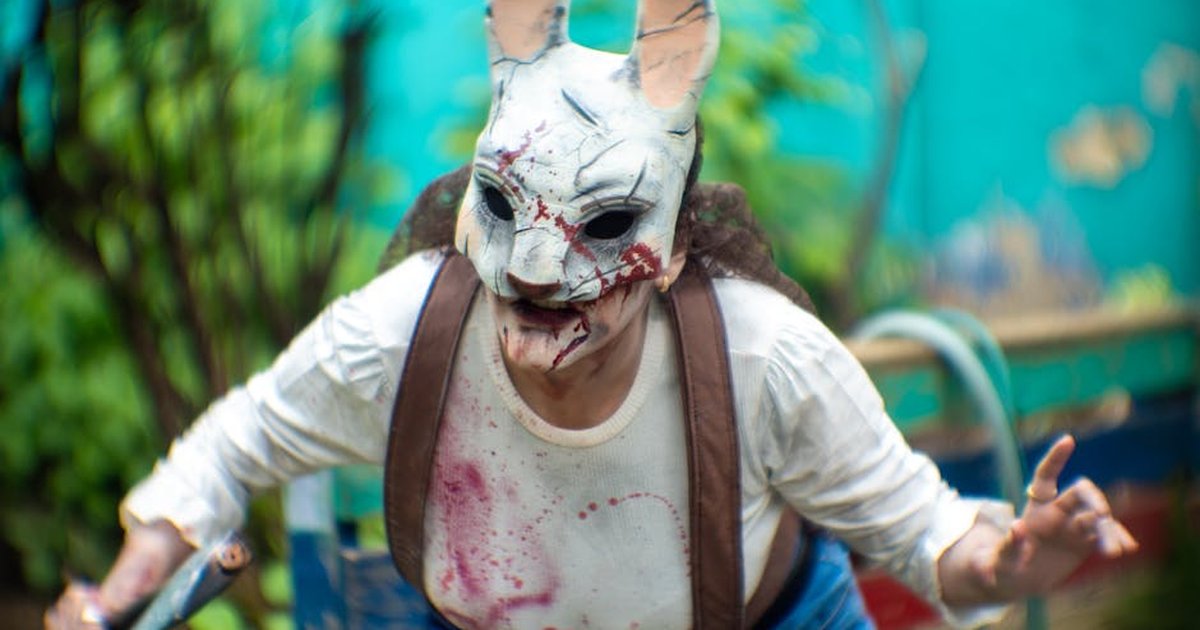 A person in a terrifying rabbit mask performs horror cosplay outdoors amid vibrant greenery.