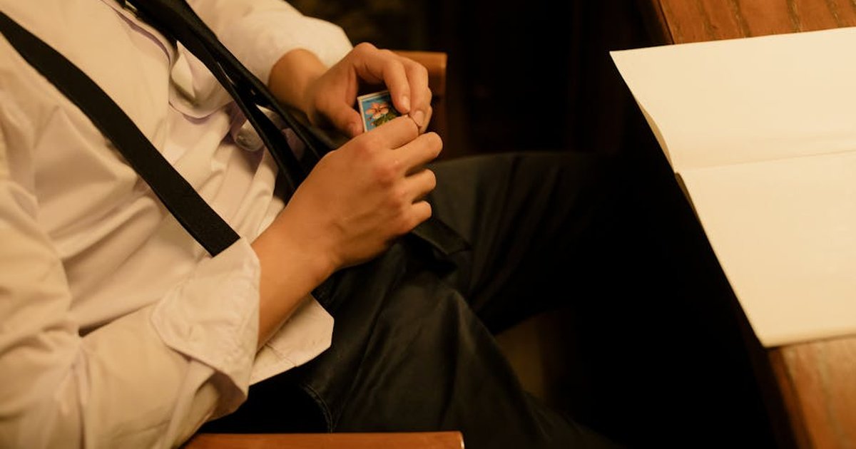 A man in suspenders holds a matchbox while sitting at a wooden desk in a dimly lit room.
