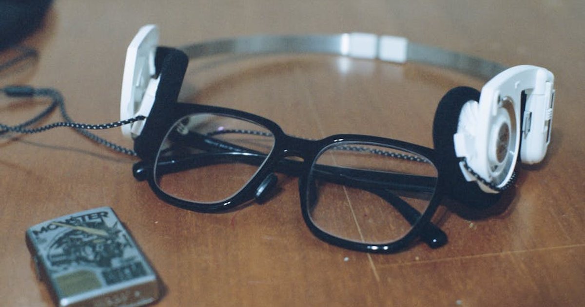 A pair of black eyeglasses and white headphones on a wooden table beside a lighter.