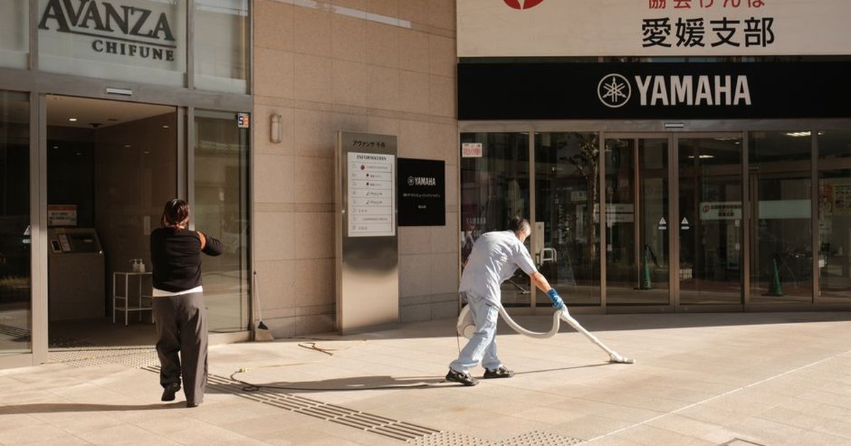 Two people clean the pavement outside a Yamaha building in Japan. Urban maintenance scene.