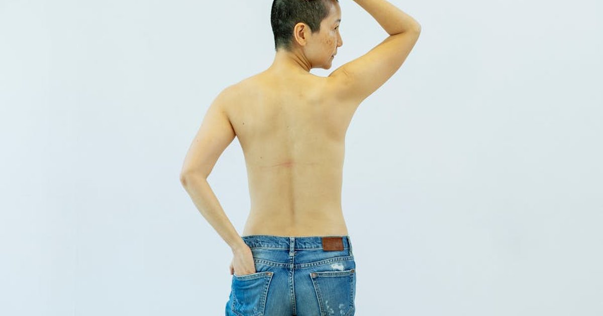 Back view of serious middle age topless Asian female in jeans with hand in pocket and on head standing in bright studio on white background