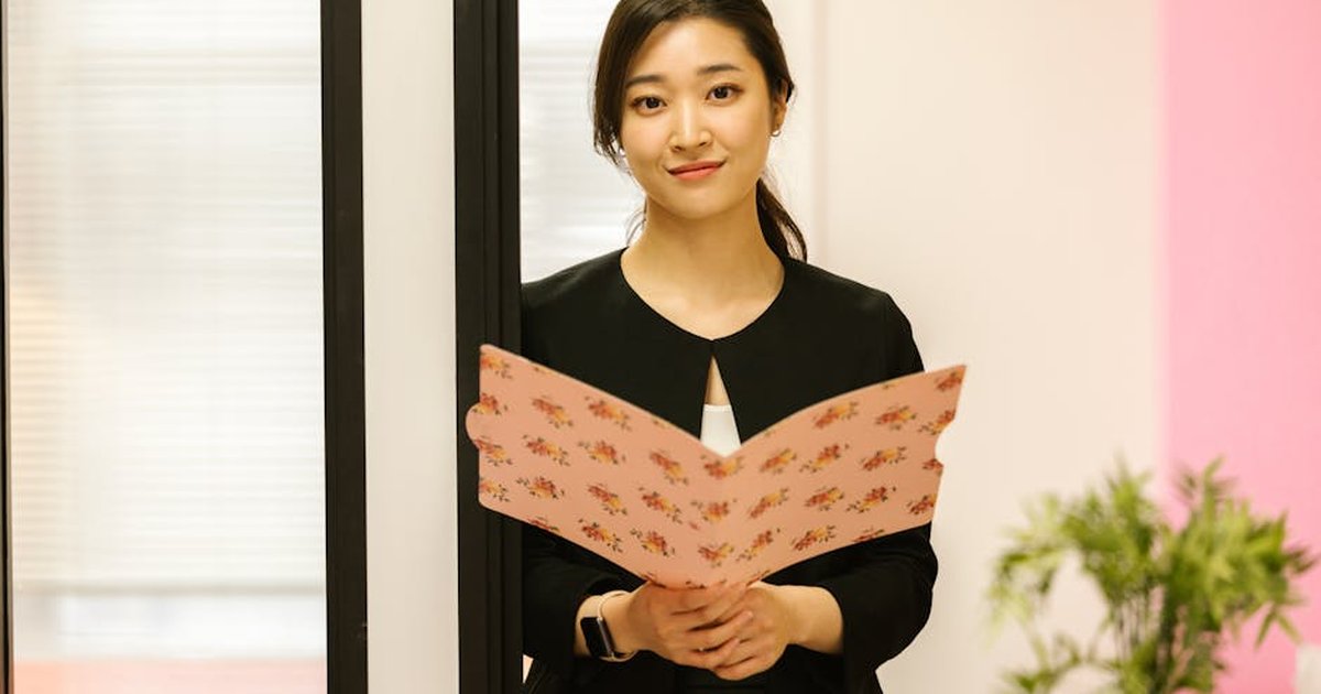 Confident woman in office attire holding a floral folder, standing in a modern workplace.