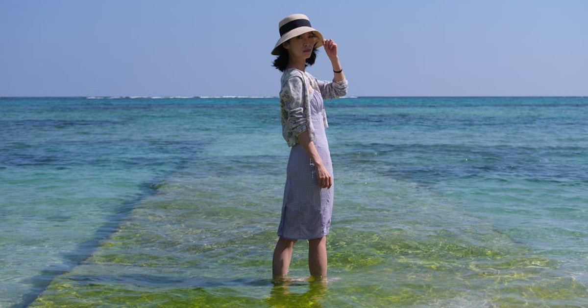 A woman stands in shallow water wearing a hat, under a clear blue sky at the beach.