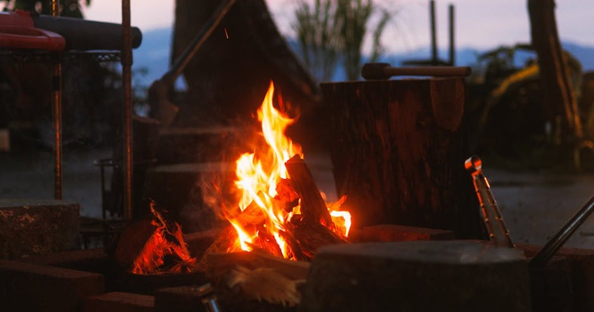 Warm campfire flickering outdoors at dusk, surrounded by nature and logs.