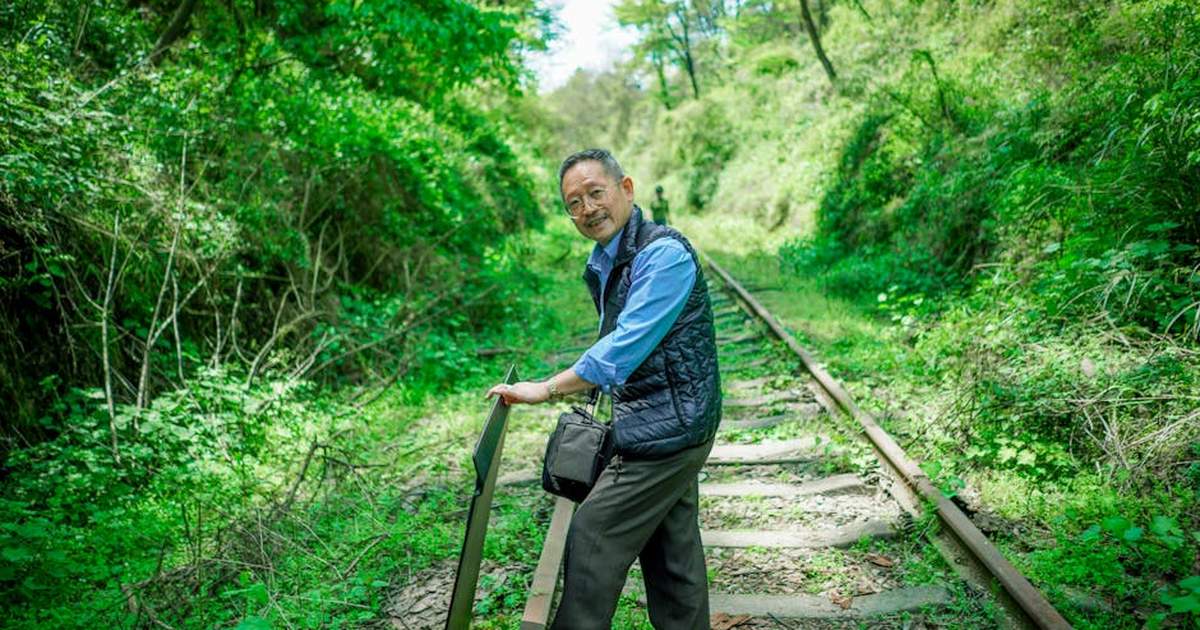 A smiling man walks along an overgrown railway track surrounded by lush greenery in spring.