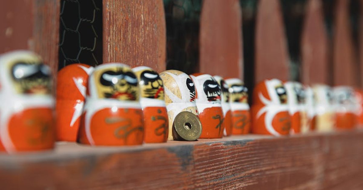 Collection of small traditional Japanese daruma dolls placed on wooden fencing on sunny street in suburb area on blurred background