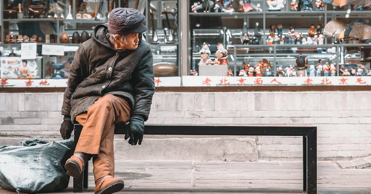 A senior man in warm clothing rests on a bench outside a Beijing shop displaying traditional items.