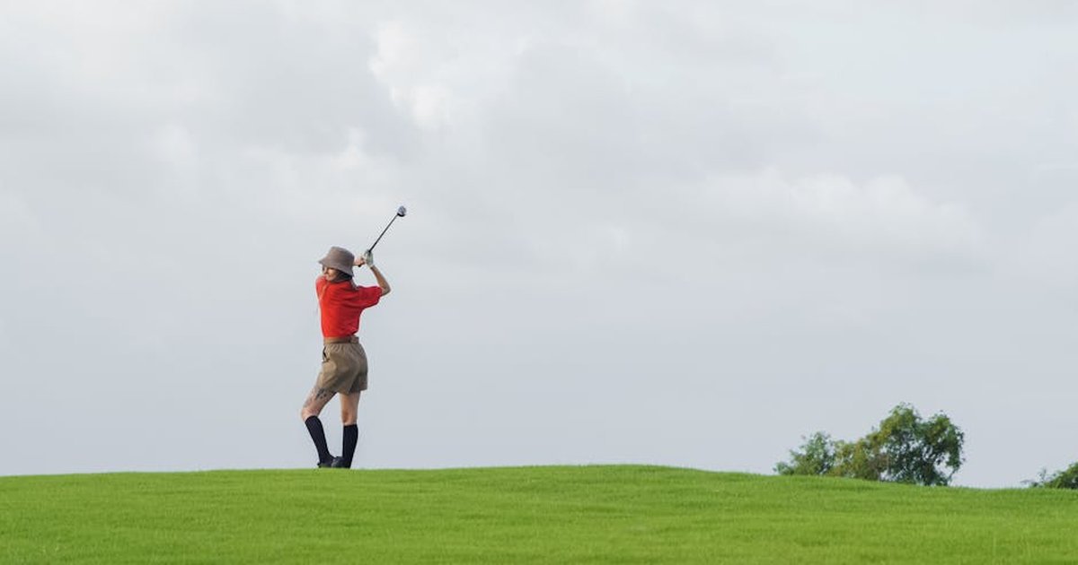 A woman in red shirt swinging a golf club on a lush green course under a cloudy sky.