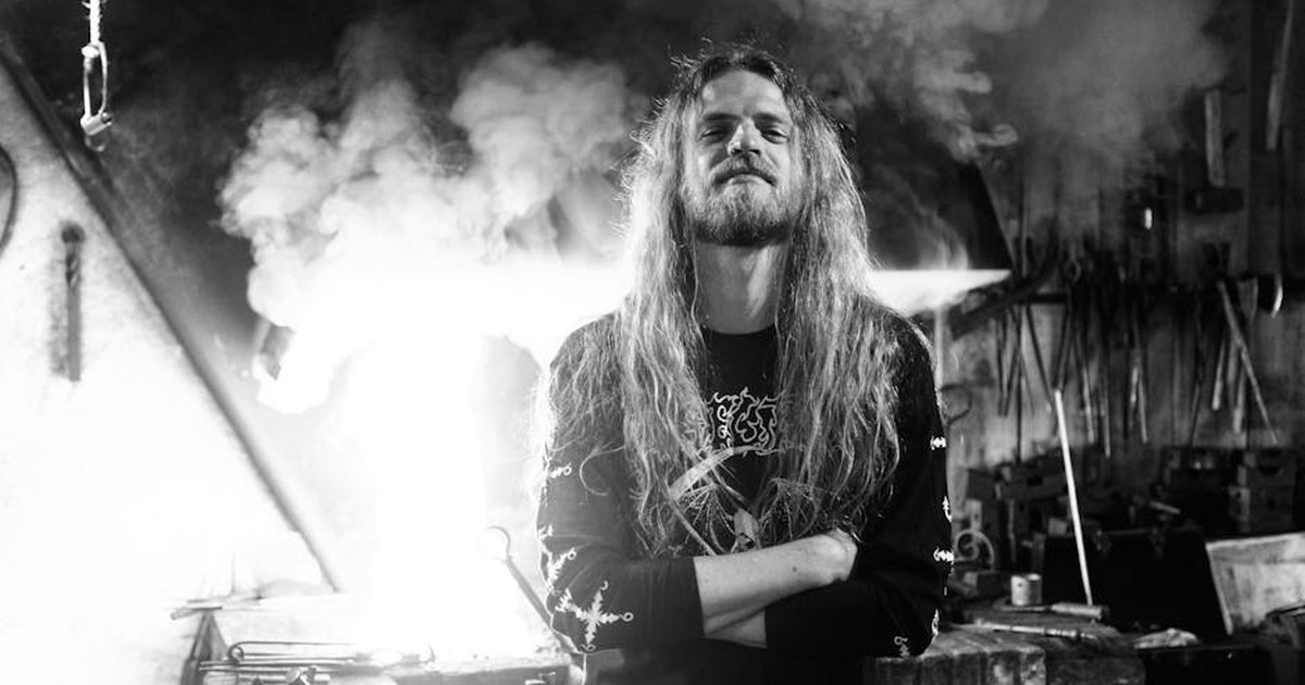 Dramatic black and white portrait of a long-haired male blacksmith in a workshop filled with smoke.