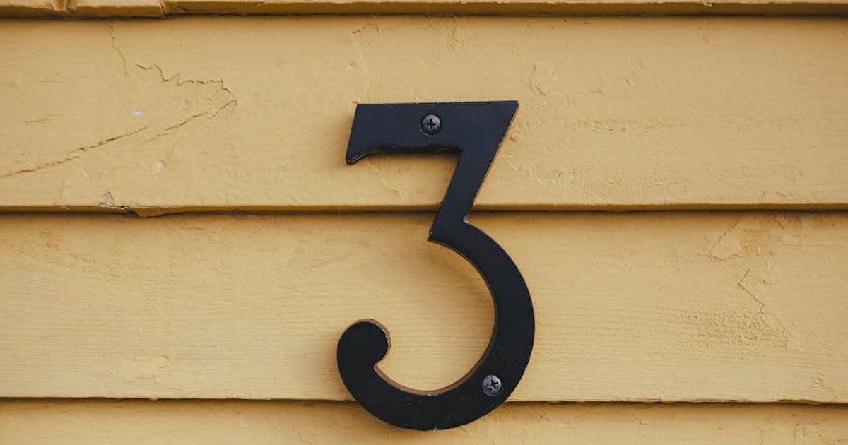 Close-up of a black number three sign mounted on a textured yellow wooden wall.