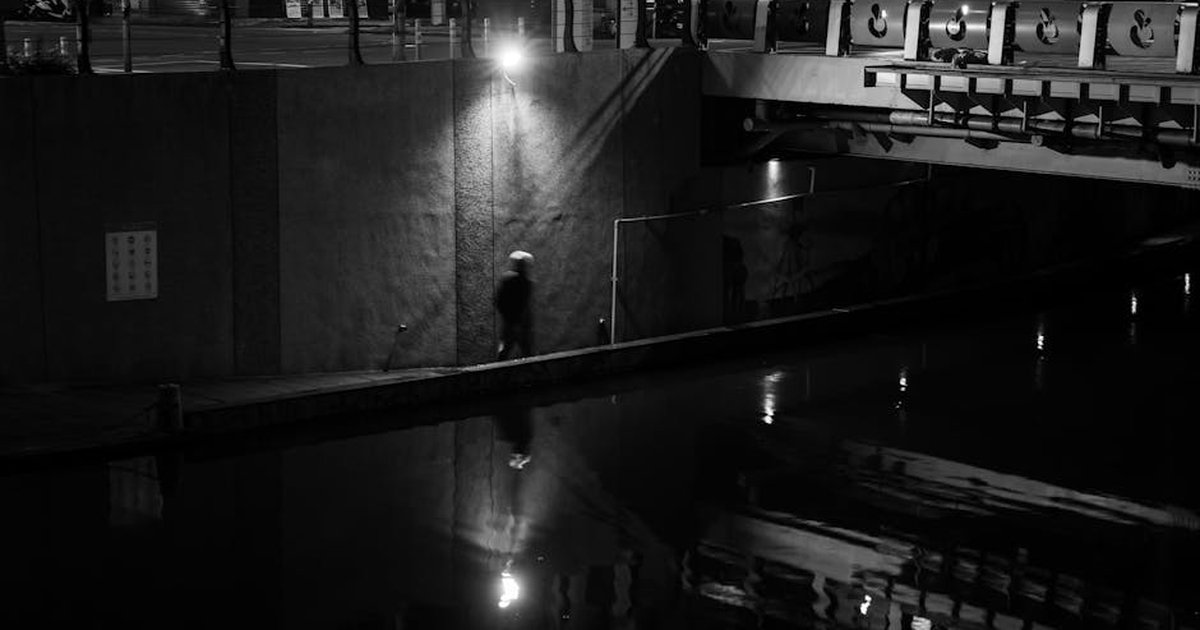 Moody urban night scene of a lone walker by a quiet waterfront, captured in black and white.