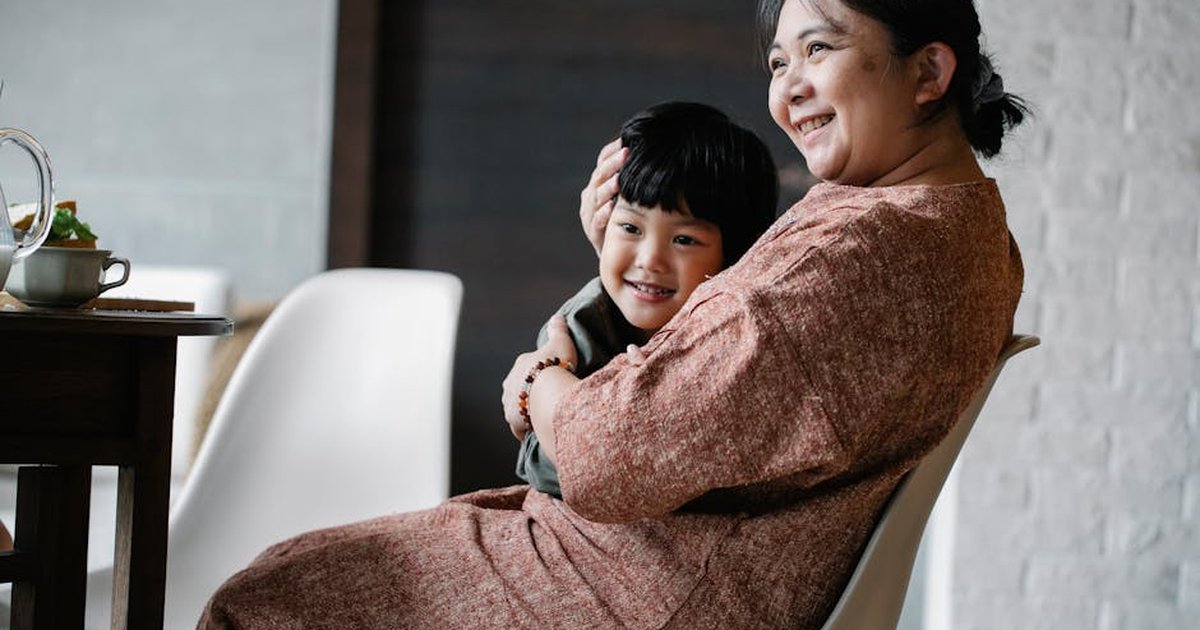 Cheerful grandmother in casual clothes sitting on chair at table while embracing little Asian grandson in kitchen with white walls