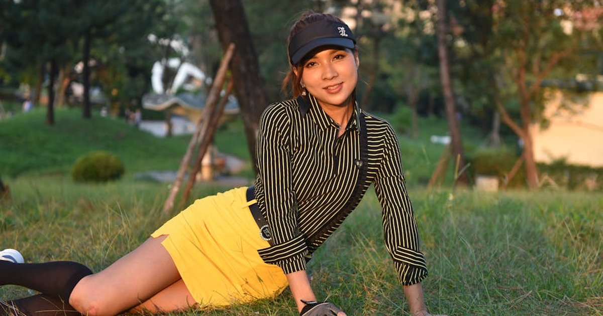 Young woman in a striped top and yellow skirt relaxing on grass amidst a sunny park setting.