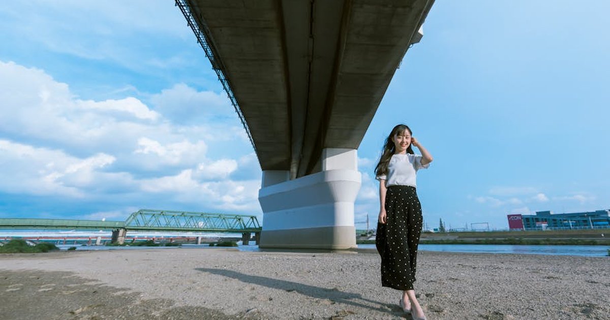 A young Asian woman smiling while standing under a large city bridge on a sunny day.