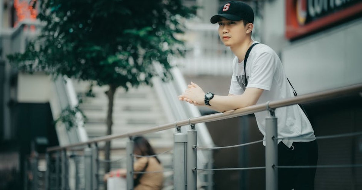 Young man in casual attire leans on railings outdoors, looking thoughtful.