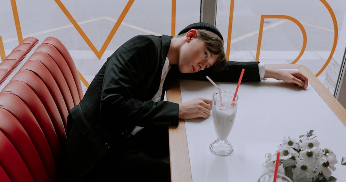 Young man in stylish outfit relaxing at a cafe booth with milkshake and white flowers.