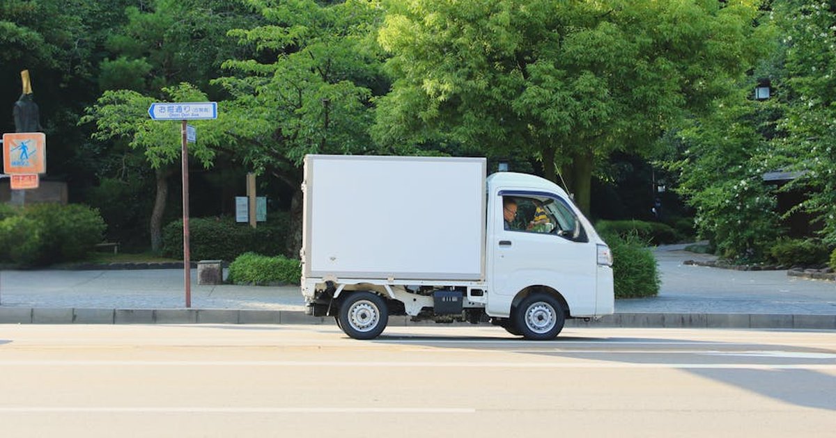 White delivery truck driving on a sunny day in Hiroshima, Japan, surrounded by green trees.
