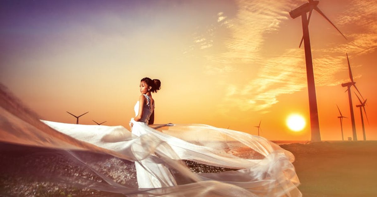 A woman in a flowing dress stands gracefully against a scenic wind farm at sunset, blending elegance with nature.
