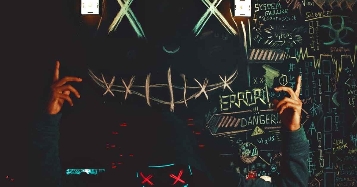 Intriguing hooded individual with neon mask against a chalkboard backdrop of cryptic symbols.