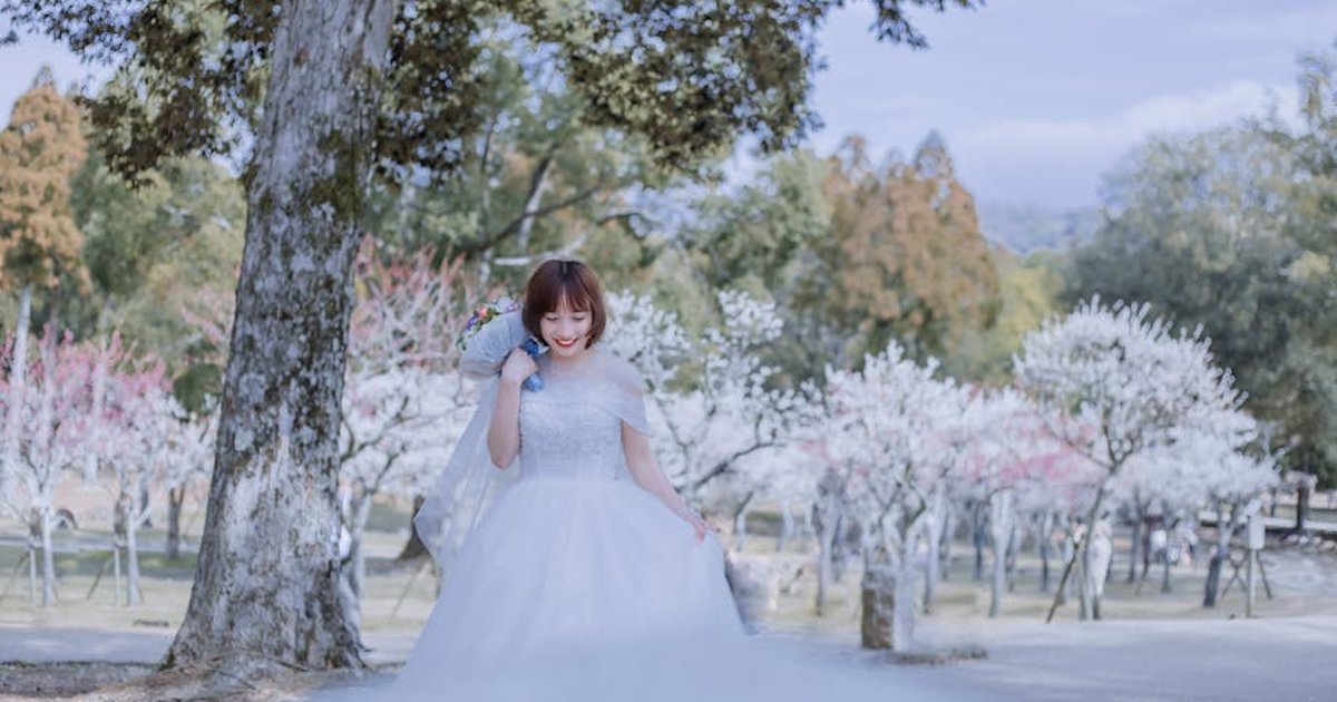 Smiling bride in a white gown holding a bouquet, surrounded by blossoming trees.