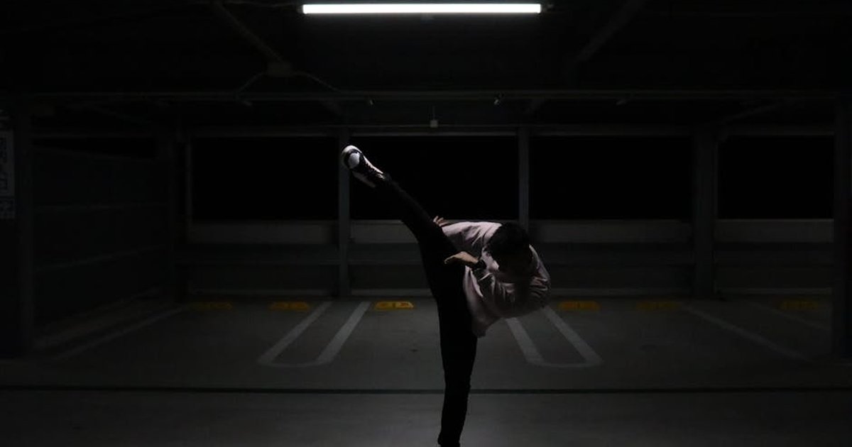 A man performs a high kick in an empty parking garage with dynamic lighting.