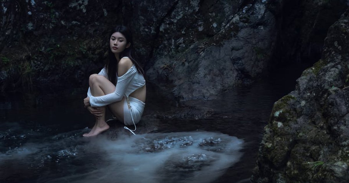 Asian woman sitting on rocks near water in a peaceful natural setting.